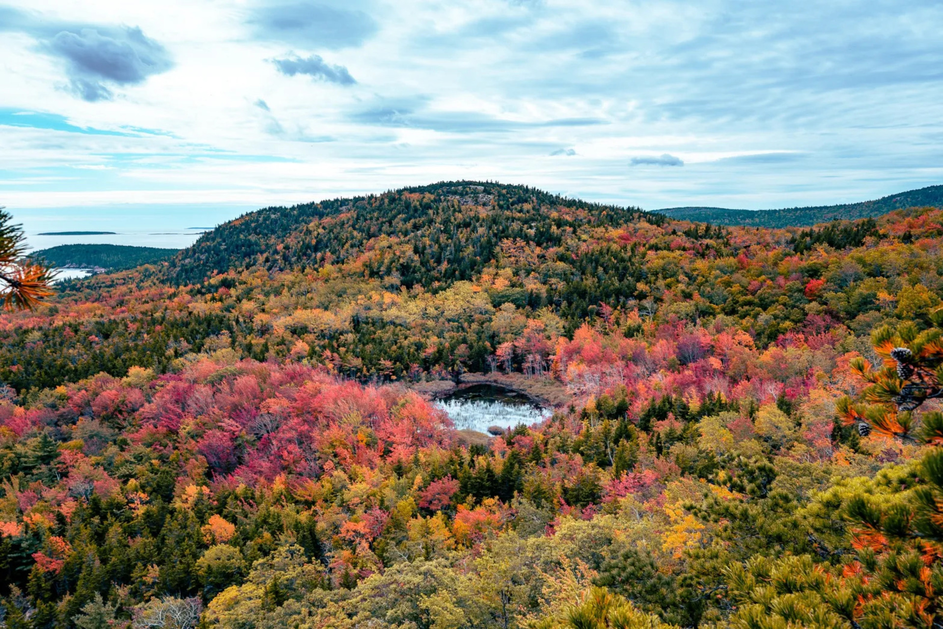 Acadia National Park's tree-covered hills are turning red and yellow under a cloudy gray-blue sky.