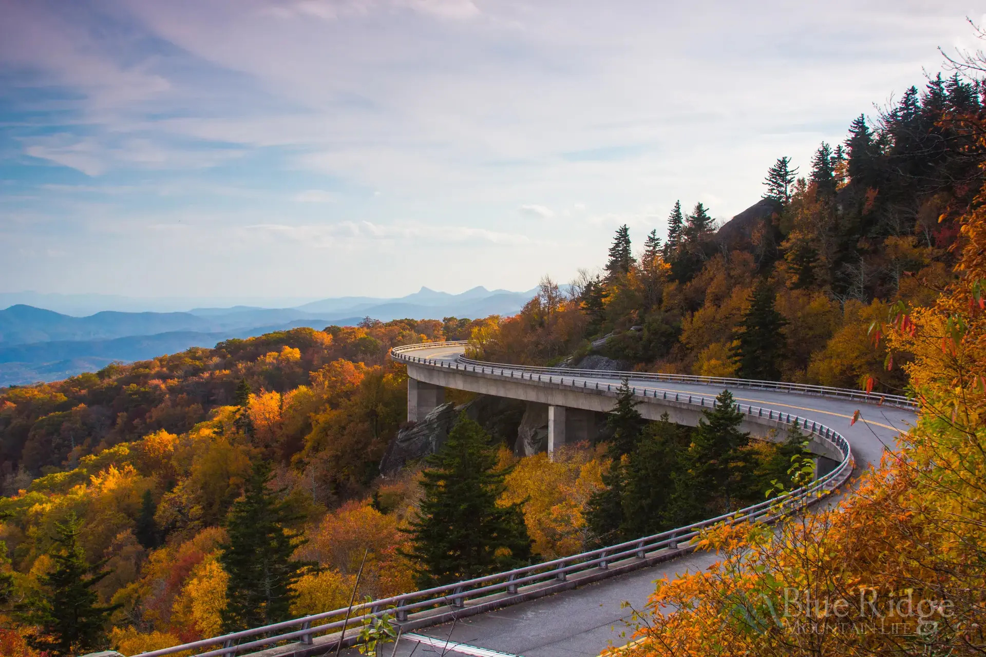 An iconic curving part of the Blue Ridge Parkway clings to the side of a mountain covered in green, orange and yellow trees. The parkway is where people have their best fall road trips.