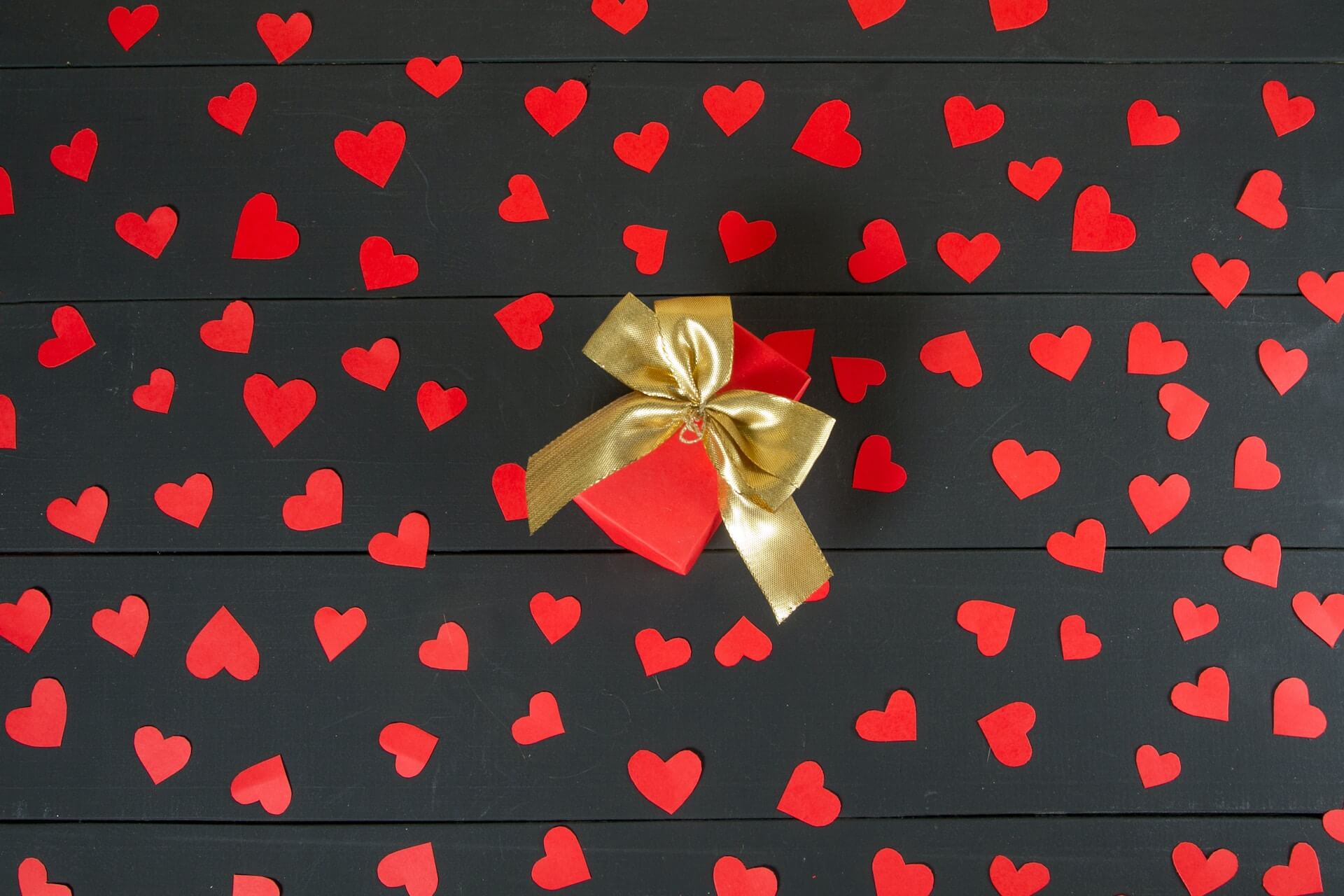 A rectangular red box sits on a floor of dark brown boards. A gold bow is tied around the box and red confetti hearts cover the floor boards.