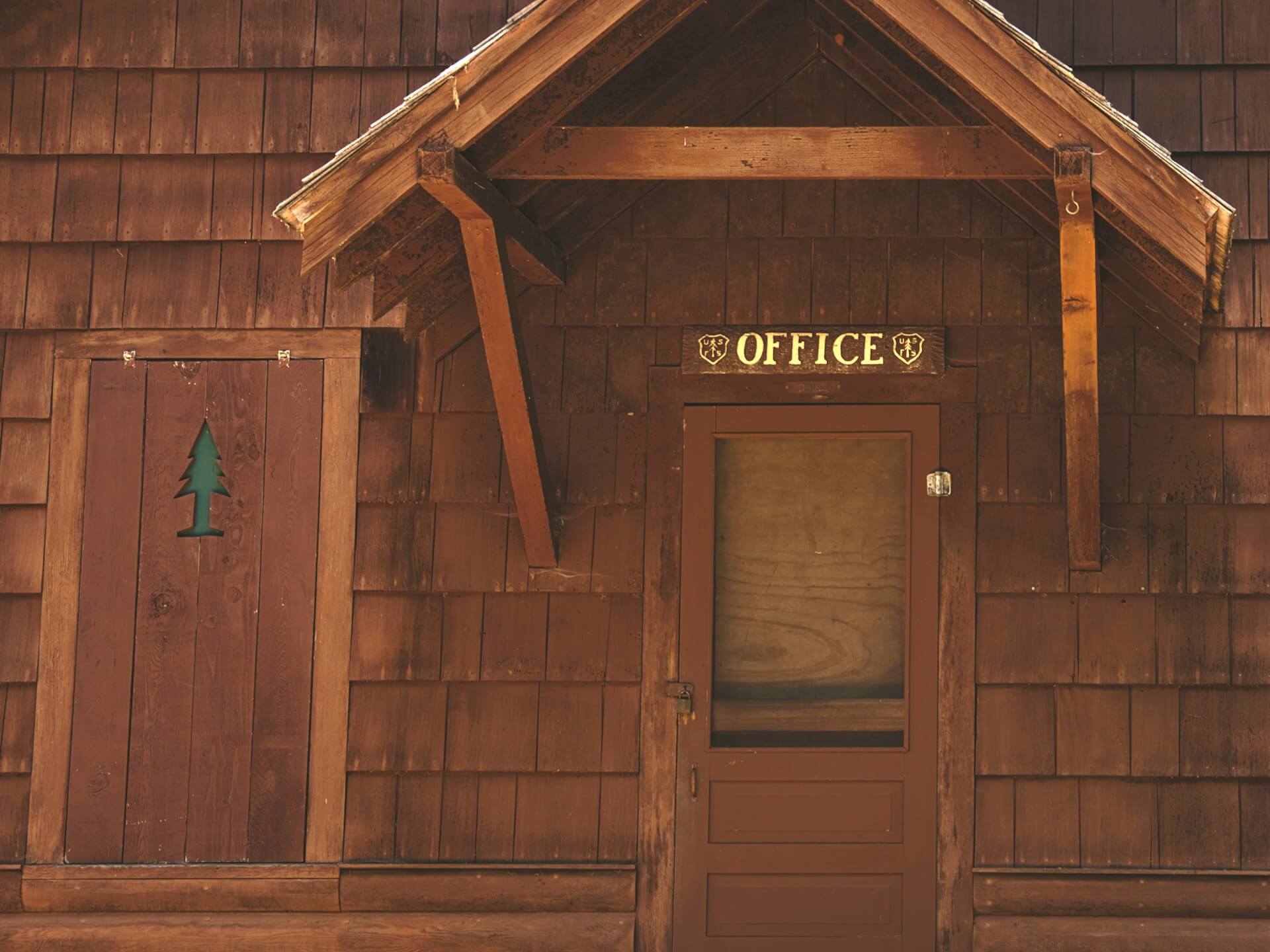 A close-up of a an office with two doors in the wooden shingle cabin. There's a sign designating the building as an office for one of the national park service jobs.