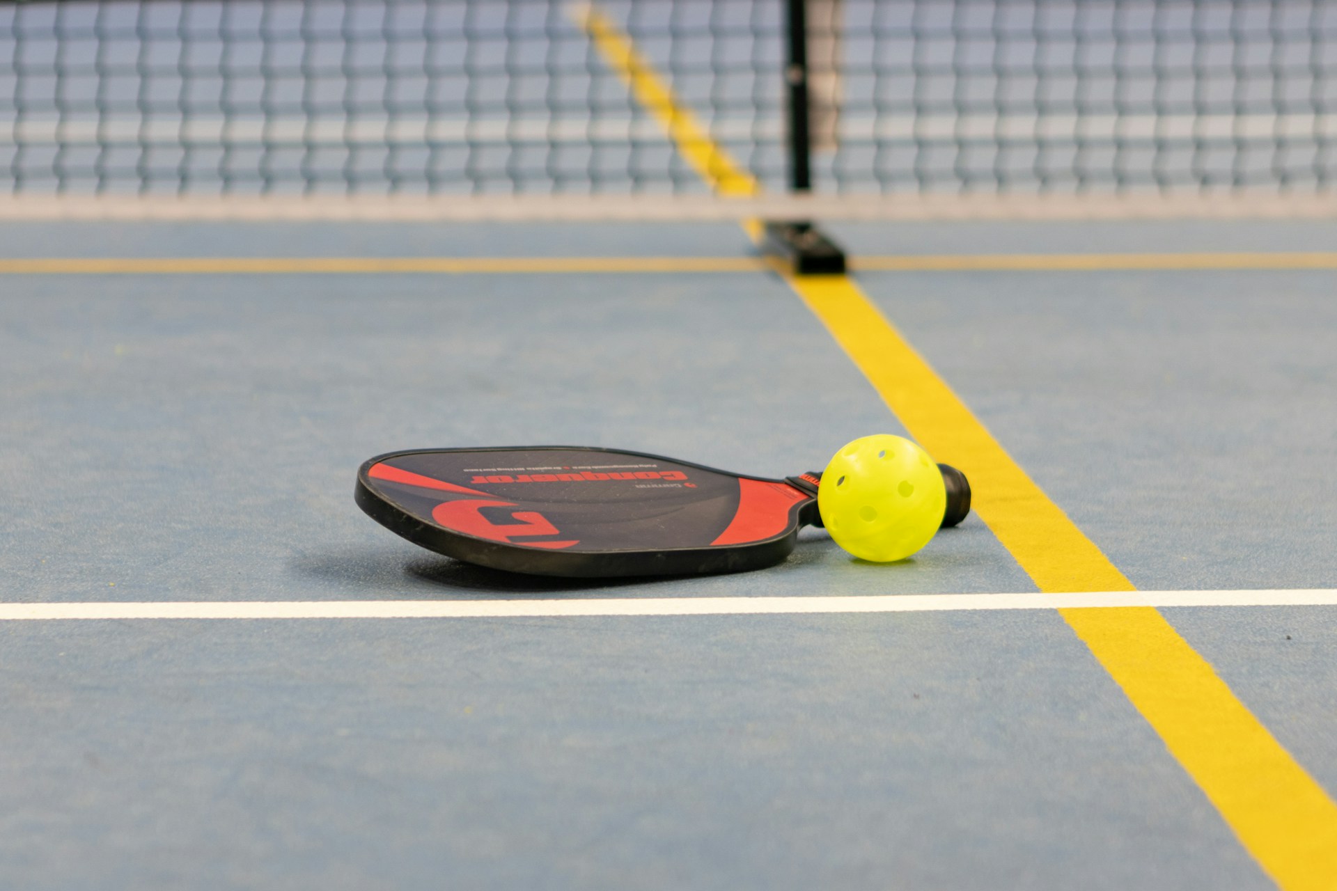 A yellow pickleball ball and red and black paddle sit on the gray, yellow and white court in front of a net.