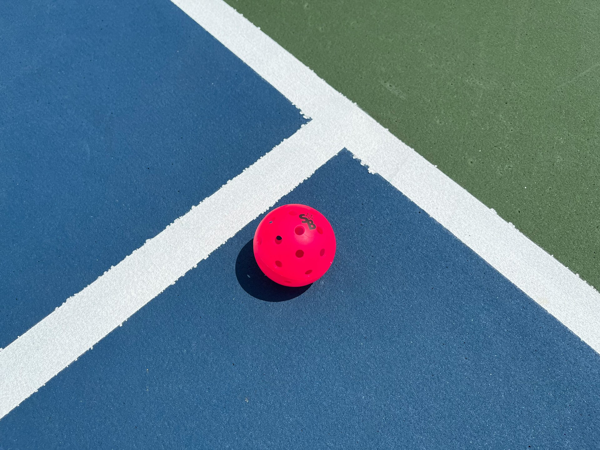 A neon pink pickleball ball sits on a blue, white and green court.