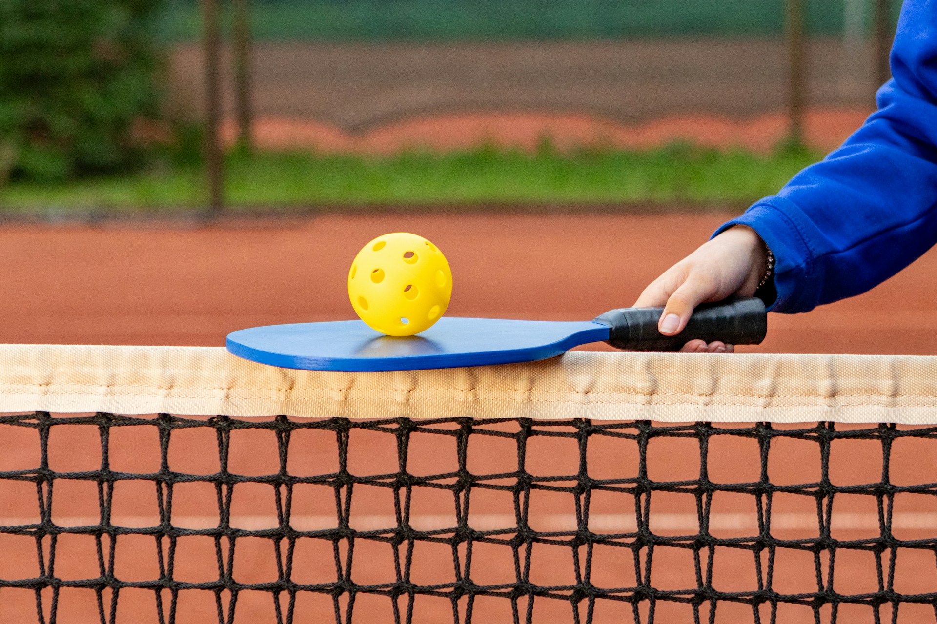 A person wearing a blue jacket holds a blue pickleball paddle with a yellow ball in the middle. The paddle rests on a net on an orange court.