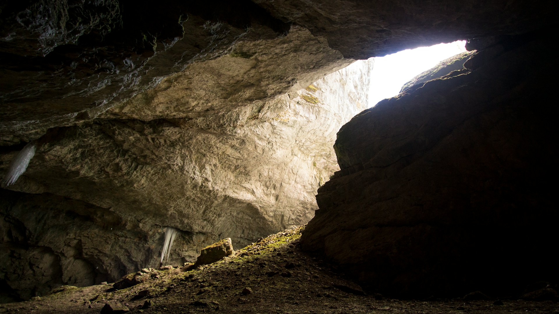The sunny entrance of a cave waits for spelunking groups in the middle of a downward sloping cavern. The rocky ground is slightly covered in moss or grass.