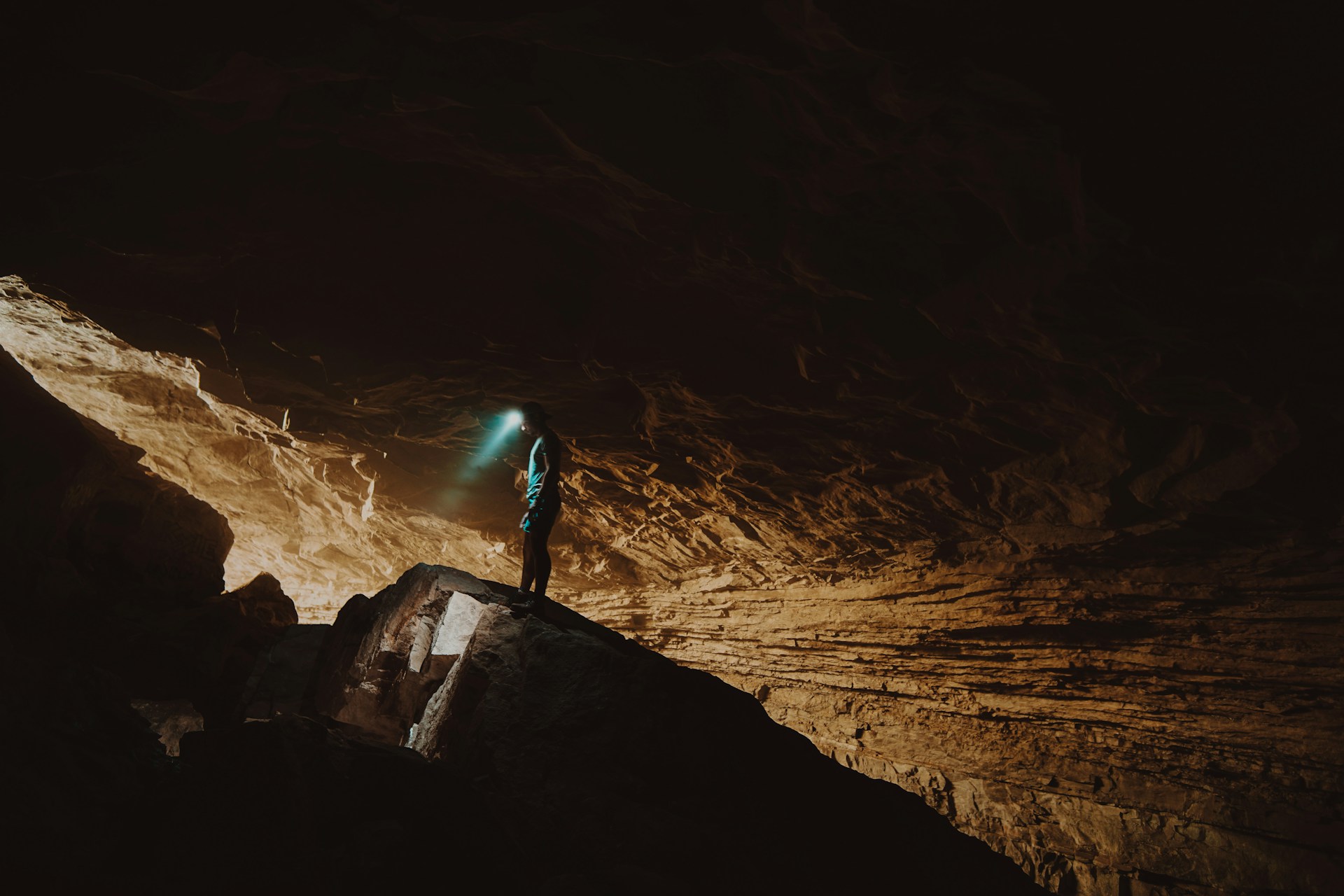 A person using a bright headlamp looks down off a cliff inside a brown stone cave. Sunlight shines from an exit far behind them.