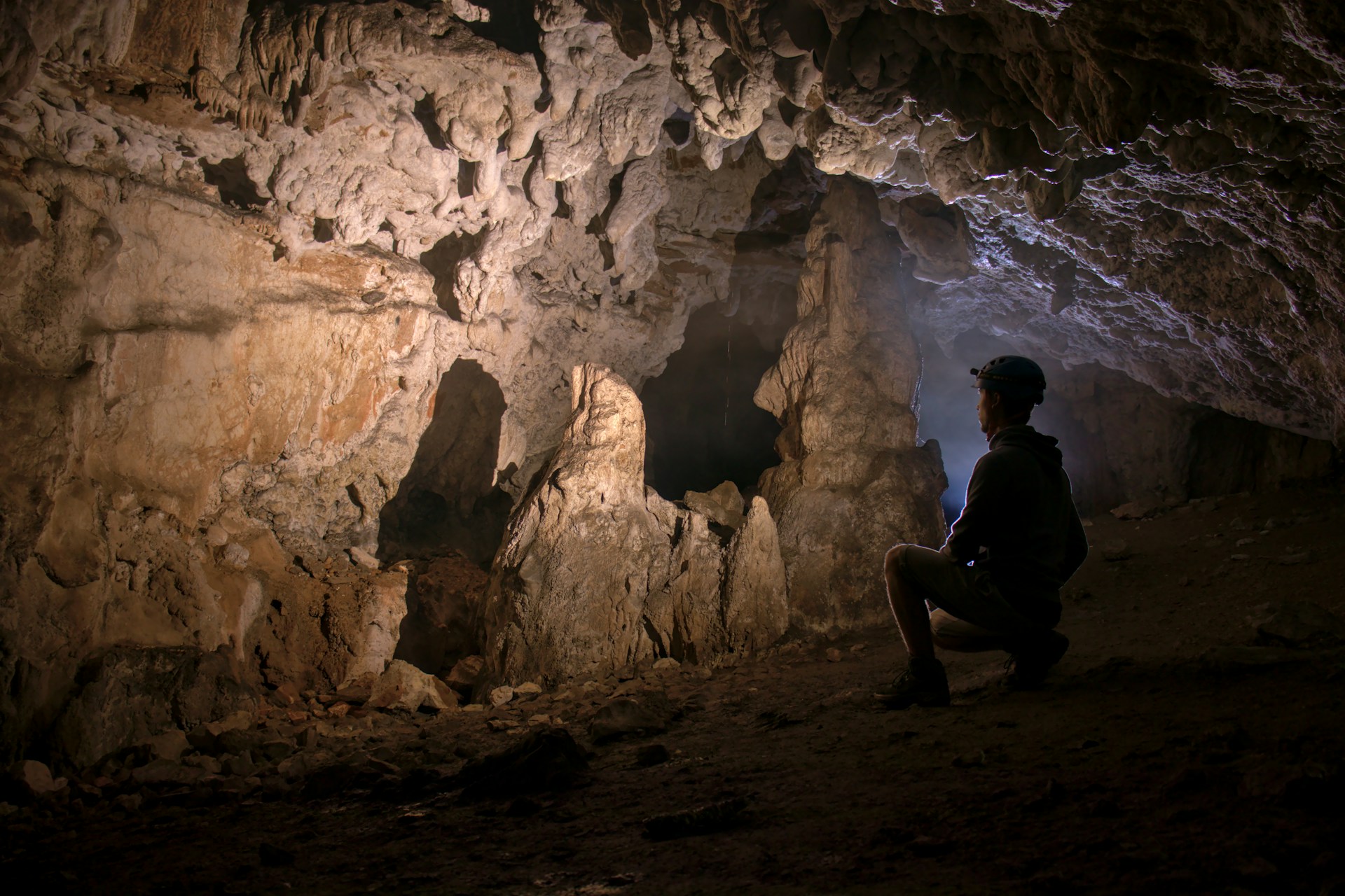 A person going spelunking uses a headlamp while kneeling to look at brown rock formations in a tunnel.