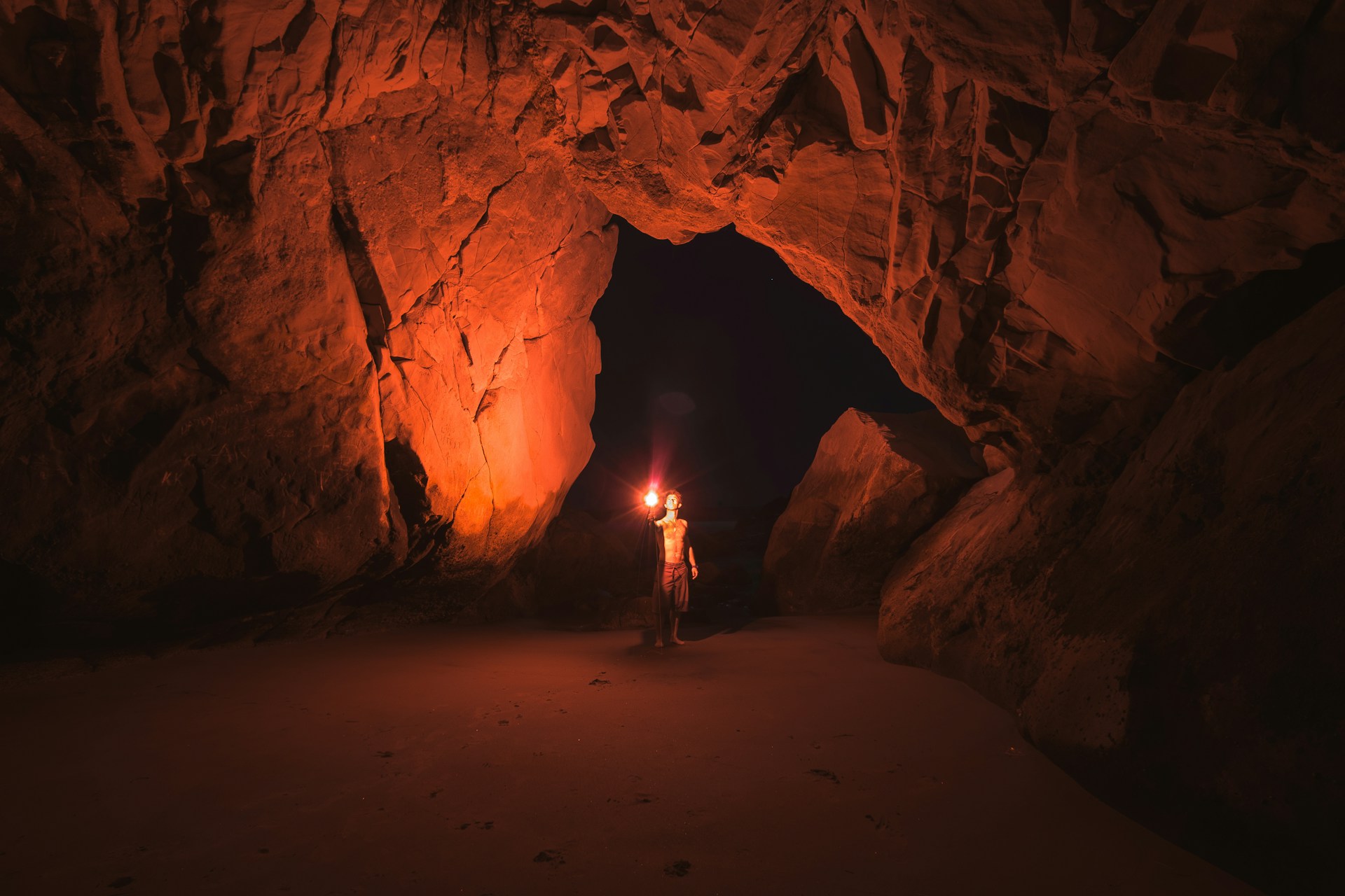 A shirtless guy wearing knee-length shorts holds a red flare. It shines red light over a tall stone cave. The floor is just sand. A large cave exit is just behind the man.