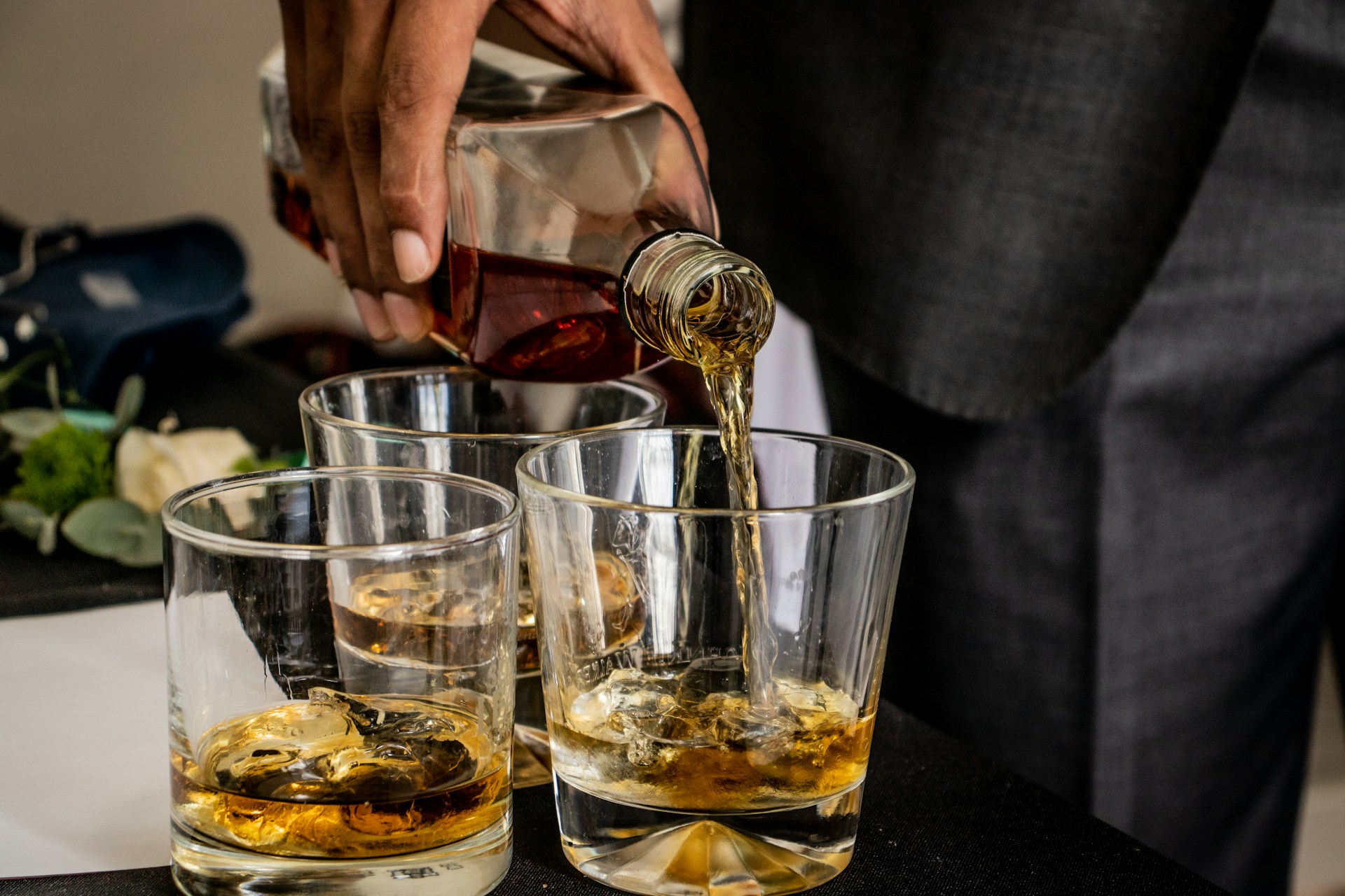 A man in a suit pours whiskey into three glasses, likely for a whiskey tasting.