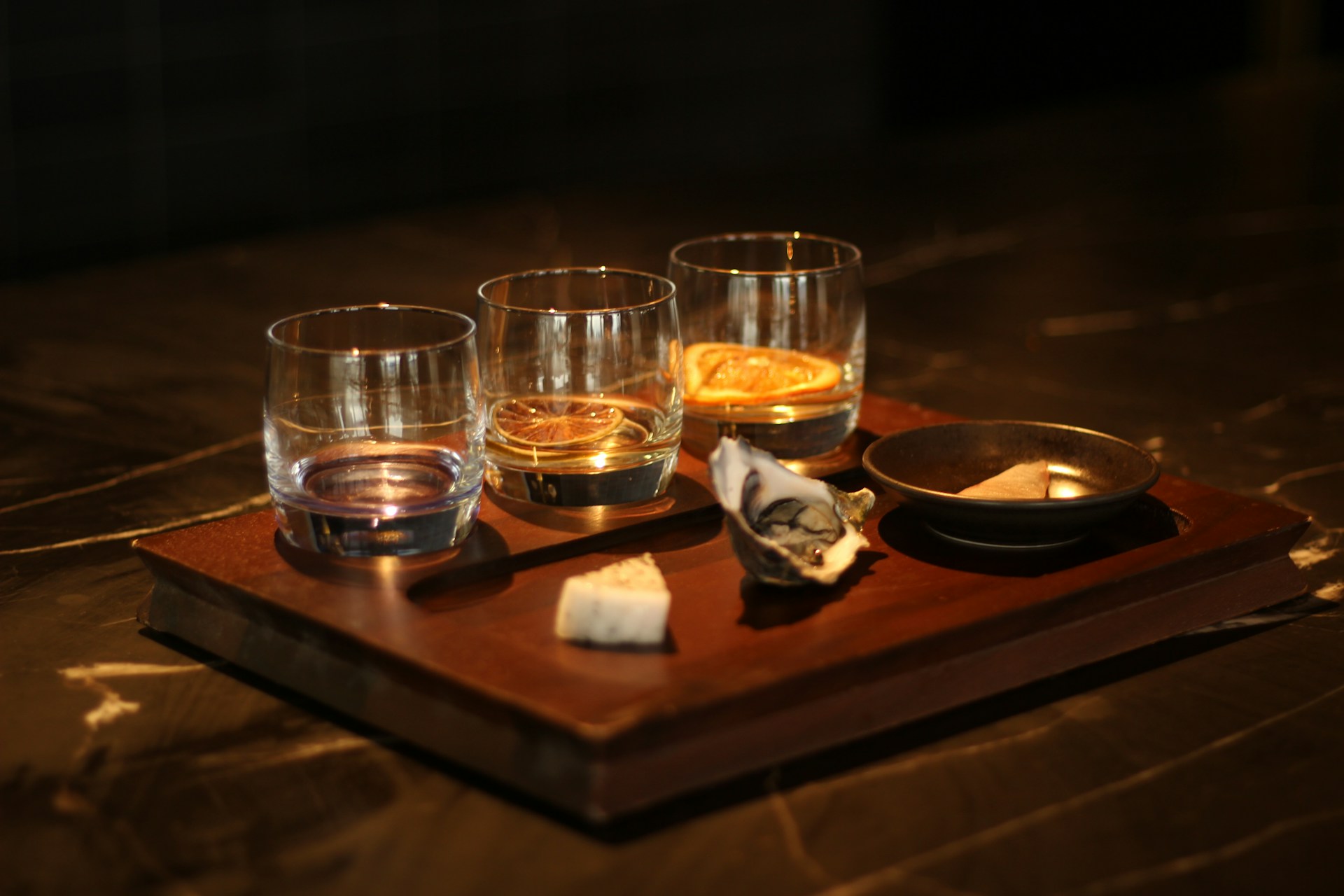 Three small glasses holding whiskey and slices of fruit sit on a serving board alongside a dish, an oyster shell and a triangle of cheese.