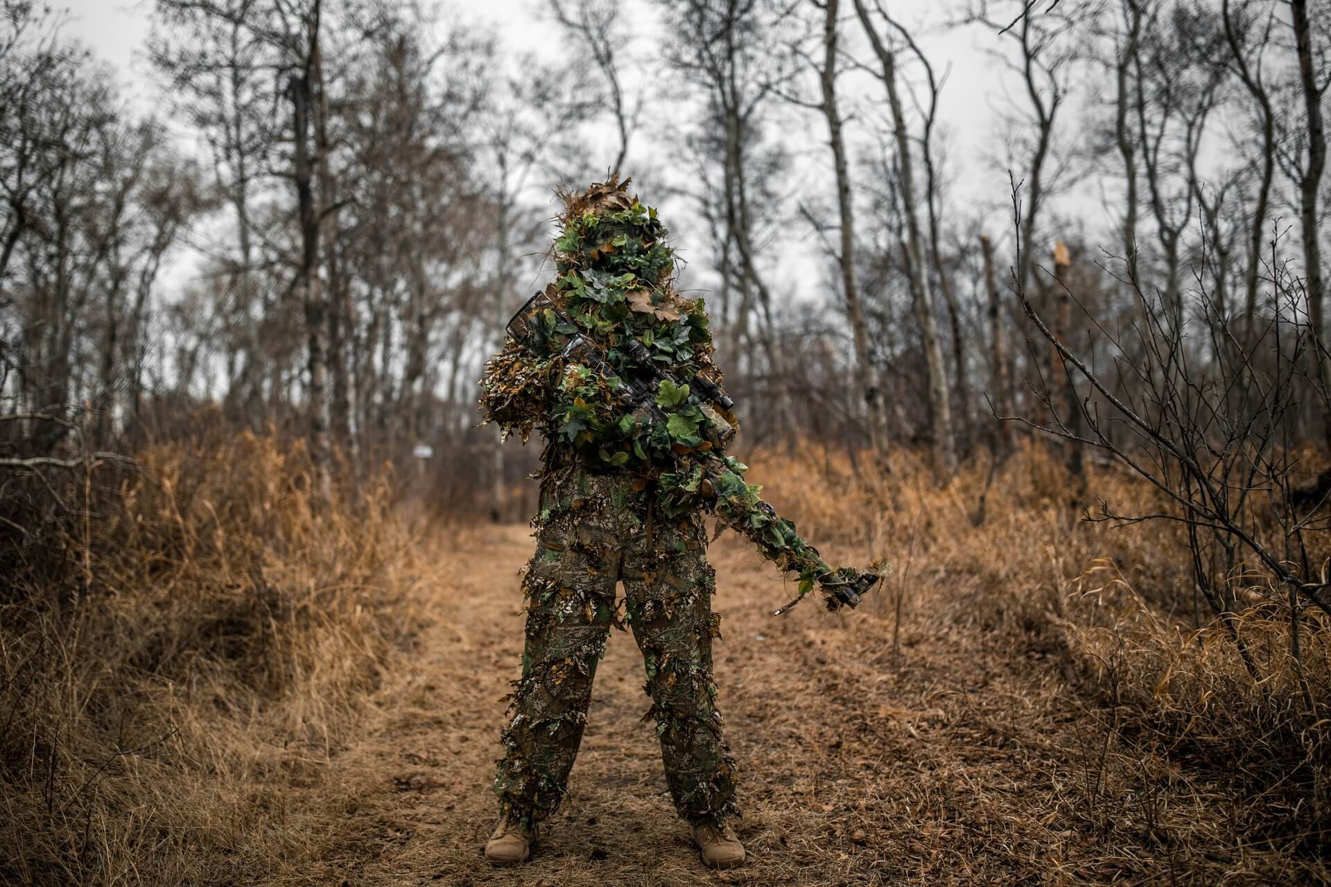 A man wearing a full-body ghillie suit while carrying a camouflaged rifle.