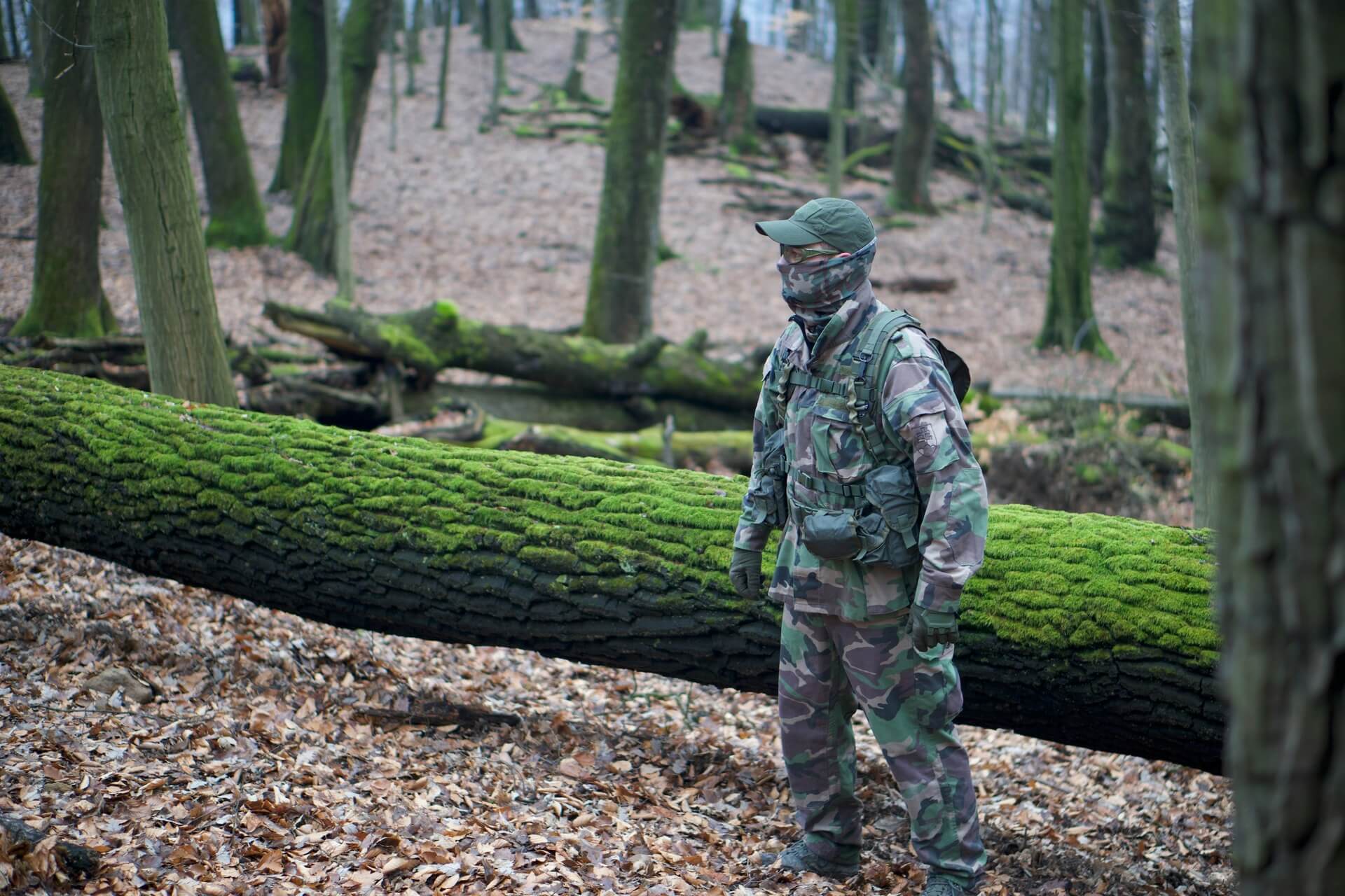 A man in a battle dress uniform standing in front of a mossy log and on dried leaves.