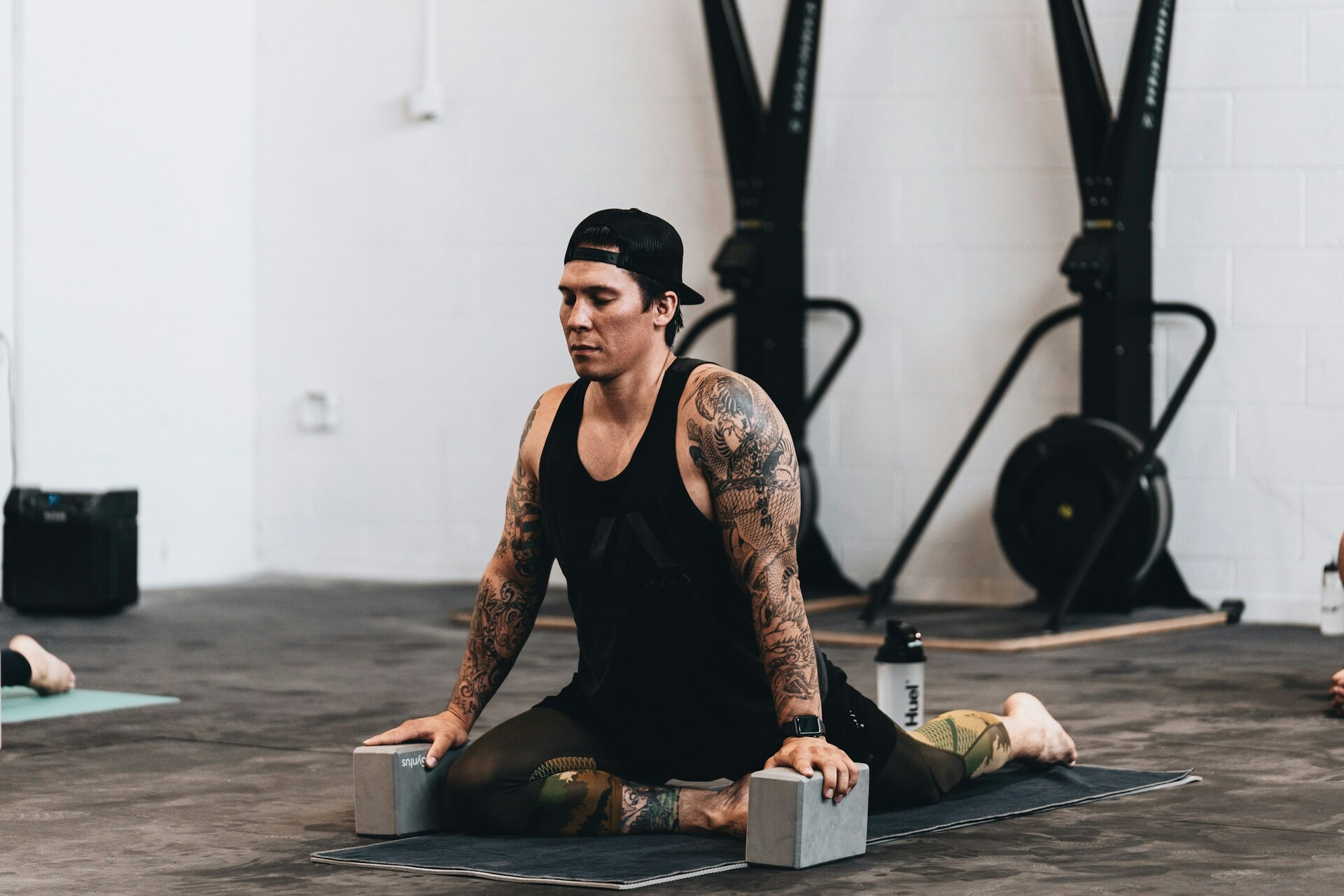 A man wearing black workout clothes and a black baseball cap stretches on a yoga mat between two gray blocks in his hands. He's surrounded by gym equipment and another person also stretching in a yoga mat. He's likely stretching because he doesn't fall for workout myths, like the idea that stretching isn't important.