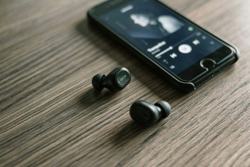 A black pair of wireless headphones and a black iPhone sit on a wooden surface. The phone is showing someone listening to a song on Spotify.
