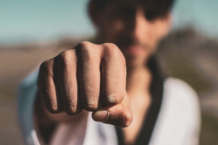 Close-up of a man's fist extended forward, with a blurred background of a clear sky and landscape. The tone is determined and focused.