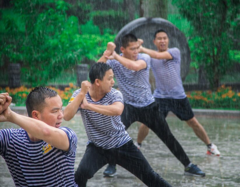 Four men in striped navy and white shirts practice martial arts in the rain. They are focused and determined, with lush greenery in the background, conveying intensity.
