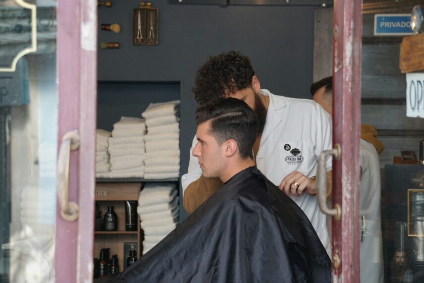 A man seated in a barber's chair while a barber trims his hair, with various hair products displayed nearby.