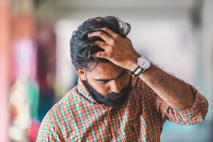 A man in a colorful orange and green plaid shirt runs his hand through his hair, looking down thoughtfully. He wears a watch, conveying a pensive, casual tone.