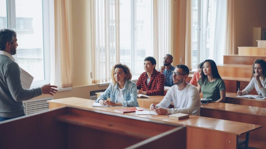 A teacher engages a diverse group of attentive students seated in a well-lit classroom with wooden desks. The atmosphere is focused and educational.