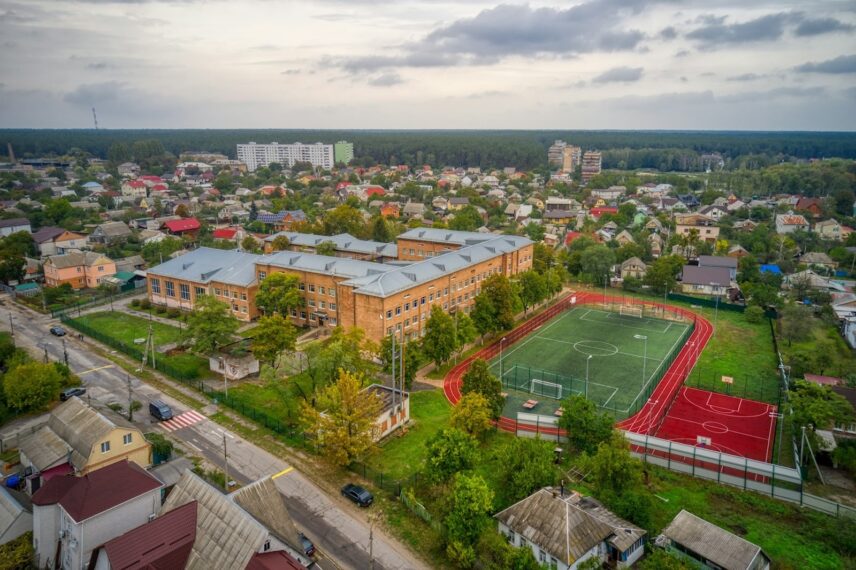 Aerial view of a large brick school building surrounded by a lush green landscape, adjacent to a fenced soccer field with a red running track. The scene conveys a peaceful suburban setting with houses and trees under a cloudy sky.