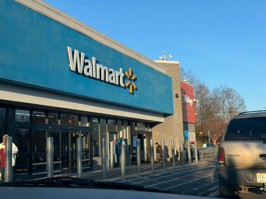 A brown Dodge car sits in front of a Walmart. The sky is blue and cloudless above. People walk in and out of the front doors.