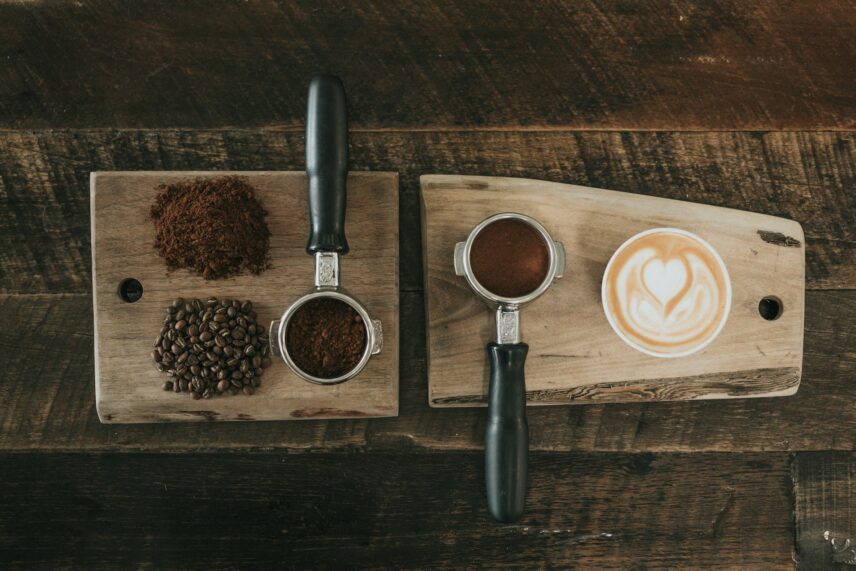 Coffee grounds sit on a wodoen surface on two cutting boards with a latte and piles of grounds and coffee beans.