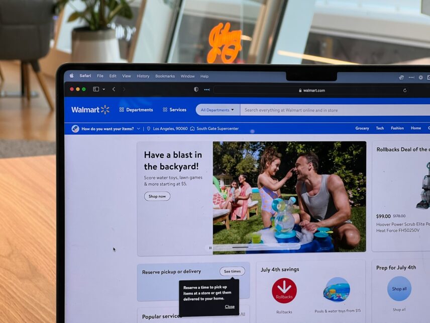 The Walmart website's homepage waits on a laptop screen. The laptop sits on a wooden table. The user is likely reading about Walmart membership benefits.
