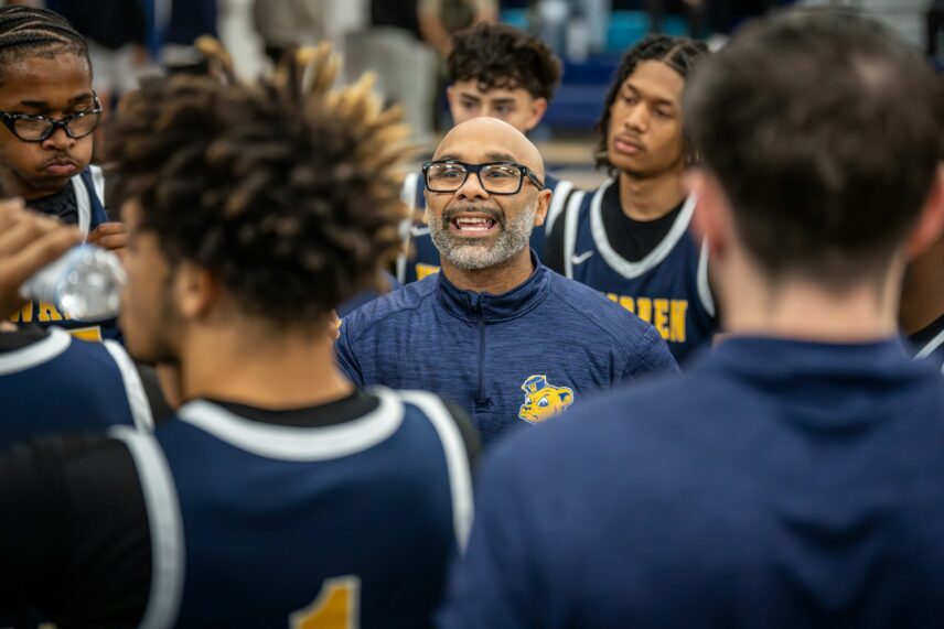 A basketball coach passionately talks to his team in a huddle; players in dark jerseys listen intently, conveying focus and motivation.