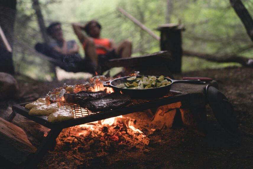 Grilled meats and vegetables cooking over a campfire. Two blurred people relax in the background, surrounded by a serene forest setting.