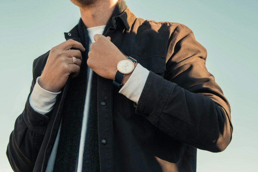 Man adjusting his dark jacket collar, wearing a white shirt and a wristwatch with a brown band, set against a clear sky, conveying a sense of style and confidence.