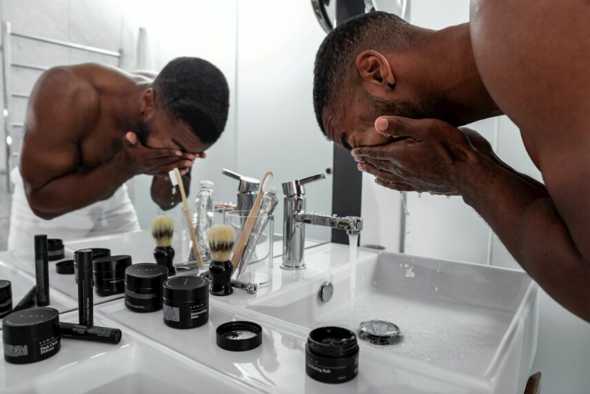 A man washes his face over a bathroom sink, surrounded by grooming products and brushes. The scene conveys self-care and a morning routine.