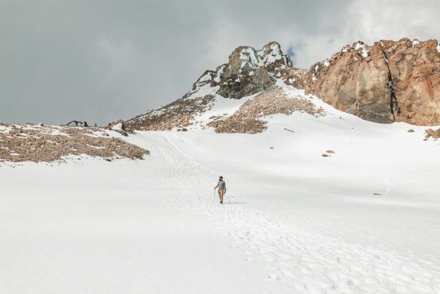 A lone hiker ascends a snow-covered slope towards rugged, rocky peaks under a cloudy sky. The scene is serene, evoking a sense of solitude and adventure.