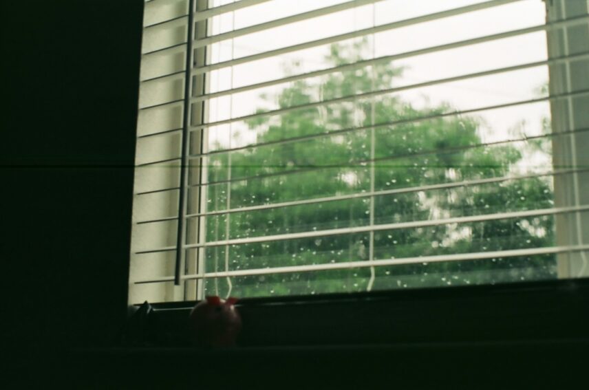 A window with closed blinds, featuring raindrops cascading down the glass surface.