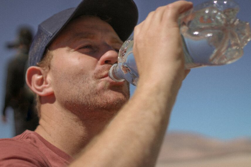 A man in a desert quenching his thirst by drinking water from a bottle under the bright sun.