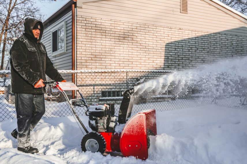 A man operates a snow blower, clearing a path through freshly fallen snow in a residential area.