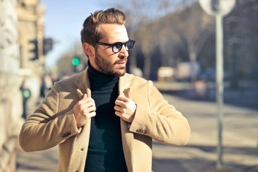 A man wearing a coat and glasses stands on a city street, looking thoughtfully into the distance.