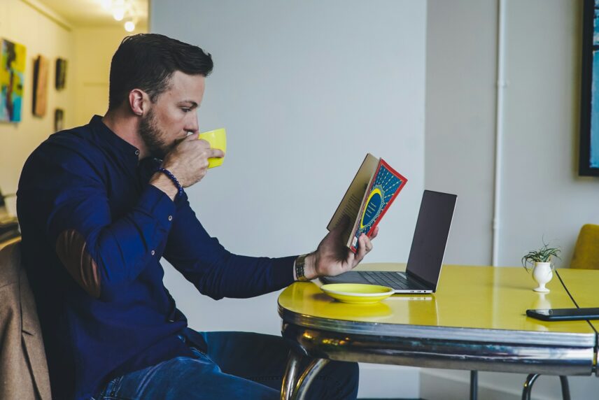 A man seated at a table and reading a book and sipping coffee, with an open laptop and plate on the table