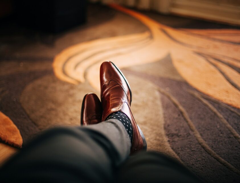 A person in brown shoes is positioned on a patterned carpet, highlighting the footwear against the floor.