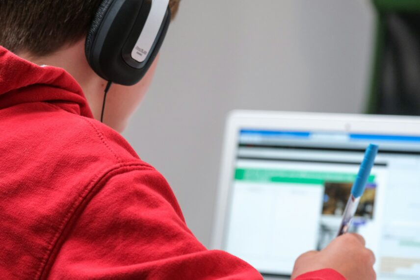 A boy in a red hoodie wearing headphones is focused on working at a computer.