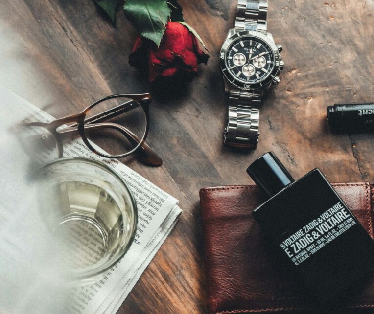 Items laid out on the table, such as a red rose, brown eyeglasses, silver watch, brown wallet, black perfume, a glass bottle and a newspaper