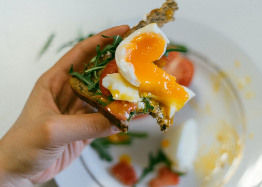 A person holding a slice of toast topped with a perfectly cooked egg, with greens and tomatoes underneath