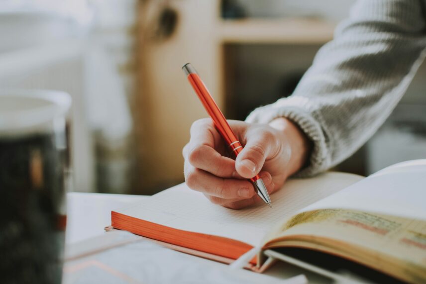 A person seated at a table, writing in a notebook with a pen, focused on their task.