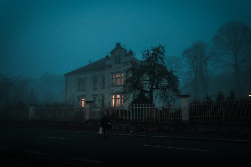 Person walking at night toward a dimly lit house through mist and fog, evoking a haunted-house atmosphere