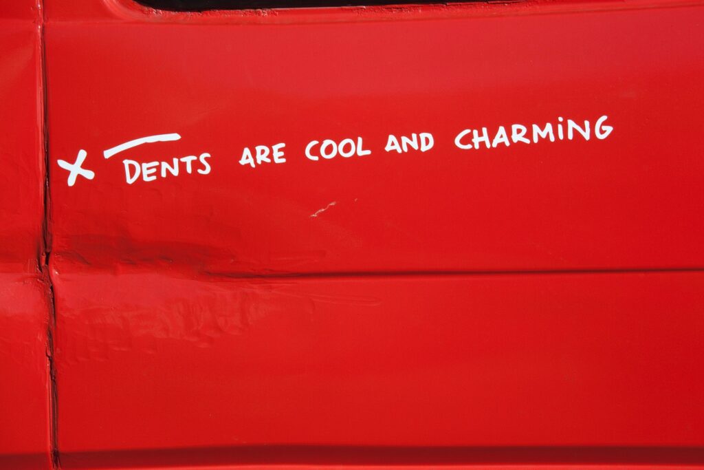 dented red car with a sticker saying "Dents are cool and charming"