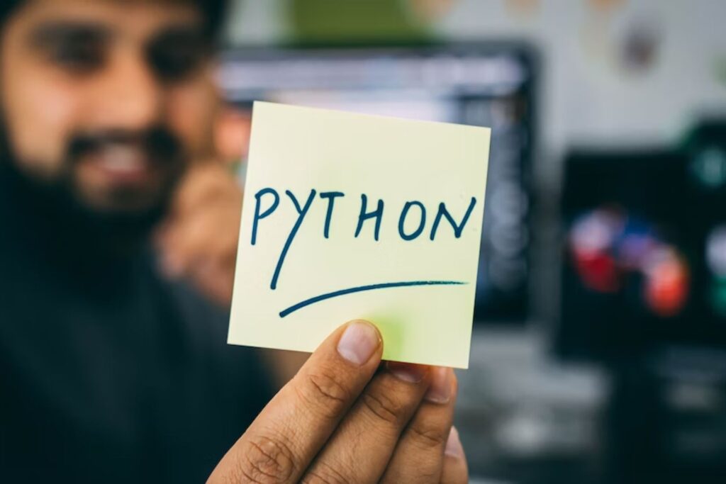 In the front focus of the camera is a yellow sticky note with "Python" written across it in green with all capital letters. It indicates that this article will teach someone how to install modules in Python specifically.