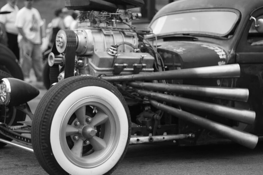 A rat rod with an exposed engine lies still as onlookers admire the car.