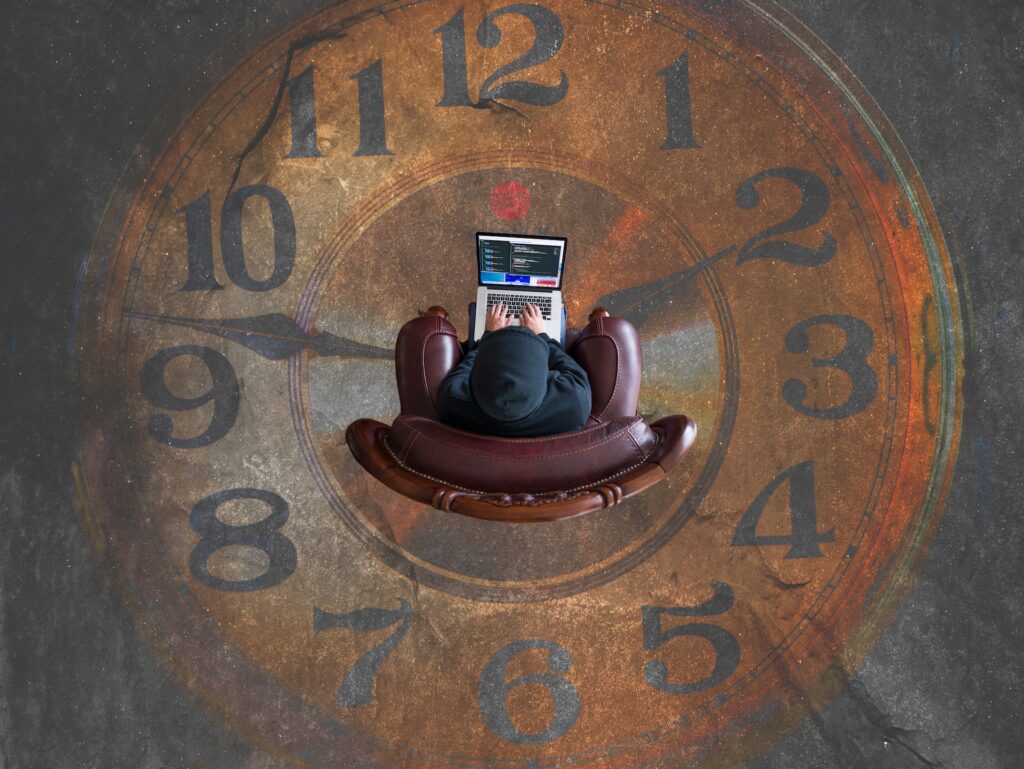 An overhead shot of a person sitting in a beanie while using a laptop. The laptop has a program running. The person's red leather chair sits in the middle of an analog clock that's part of the concrete floor. The clock reads 1:47.