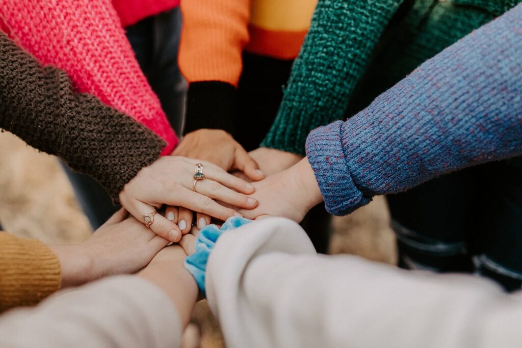 Eight people hold their hands in the center of their circle, hands on top of each other. Each person wears longs sleeves in a different color.