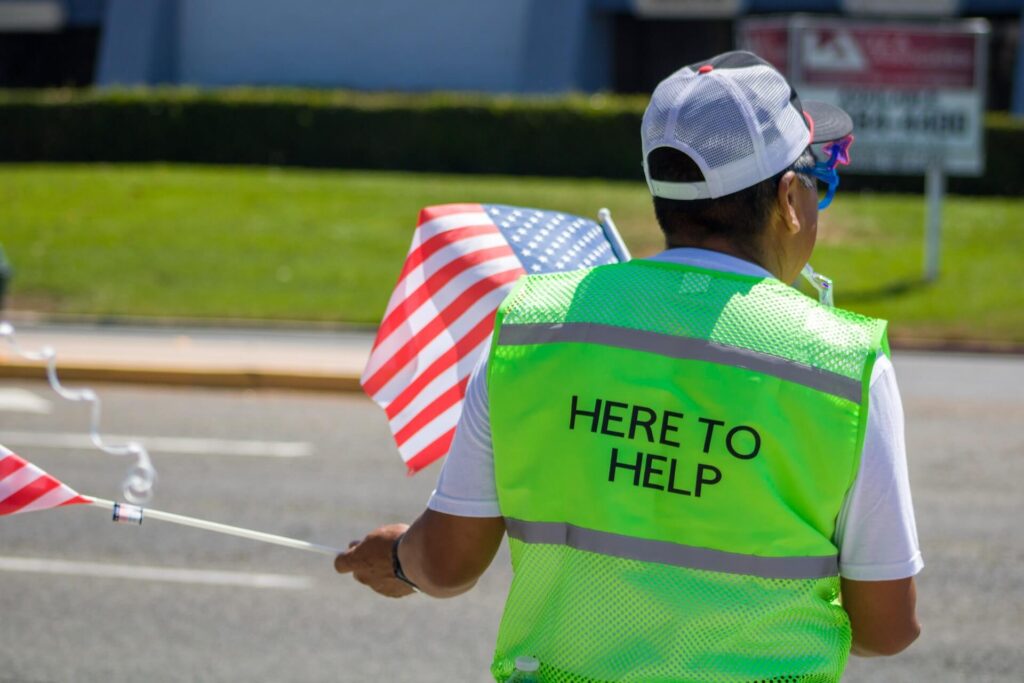 A man waving two small American flags stands facing away from the camera. He wears a green safety vest that says Here to Help. He also wears a white baseball cap and blue plastic sunglasses.