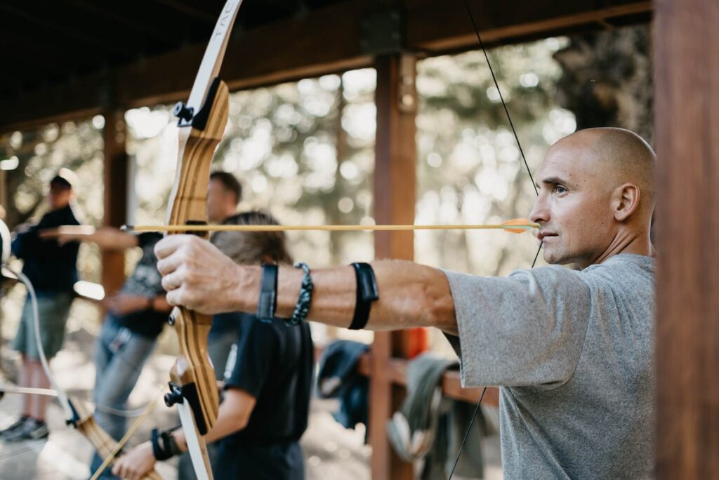 A man standing in a gray shirt with his wooden bow drawn fully back. He wears an arm brace and appears focused on a distant target. Other archers ready their bows beside him. They're at a shooting range.