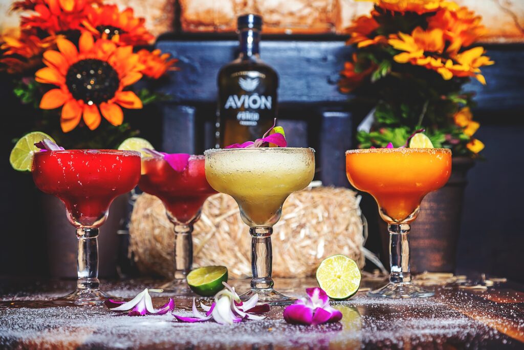 Red, white, and orange chilled margarita glasses stand on a table. Purple flower petals and lime sit around each base. Orange flowers are in the background, a dark chair, and a bottle of Avion tequila.